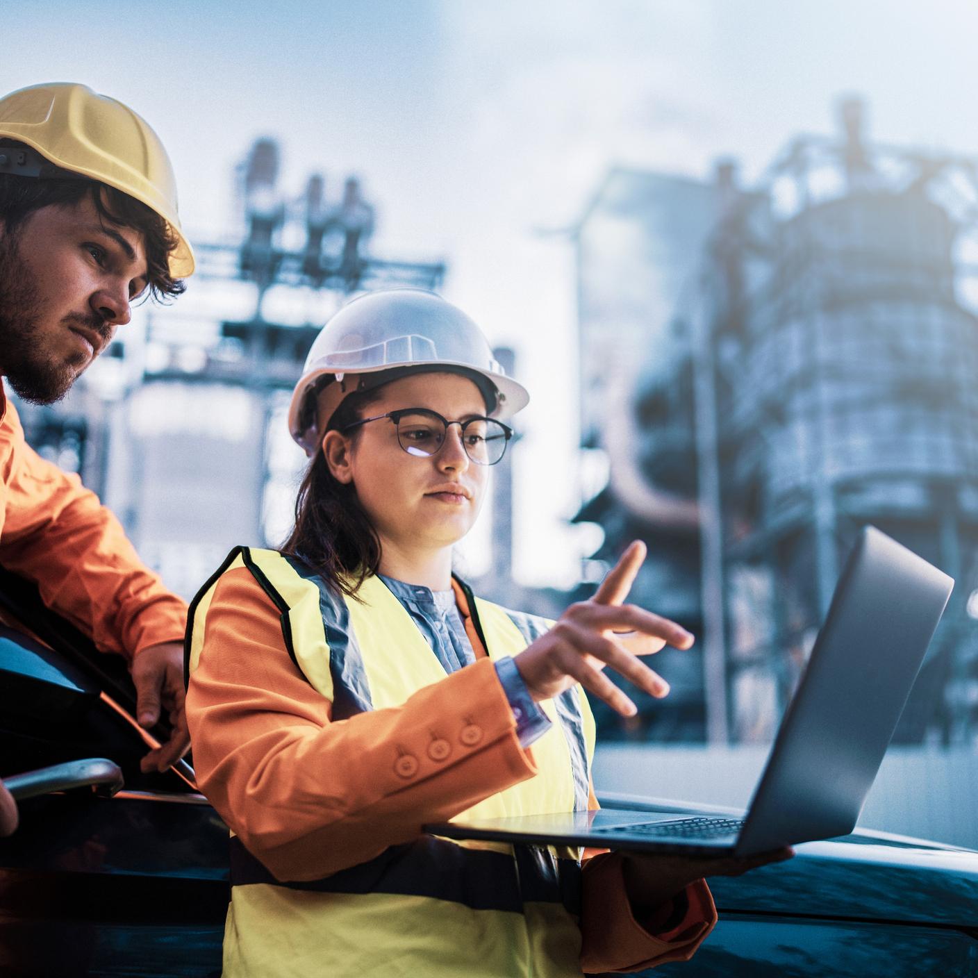 Two consultants in safety attire reviewing data  on a laptop in front of a power plant.