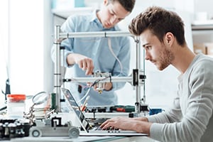 Two men working in a room, one is seated in front a of a laptop, and the other is in the background, working on a 3D printer