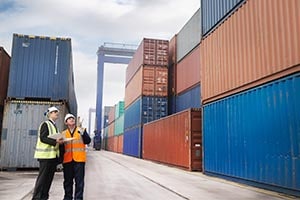 Two people wearing PPE stand in between shipping containers, and are checking containers