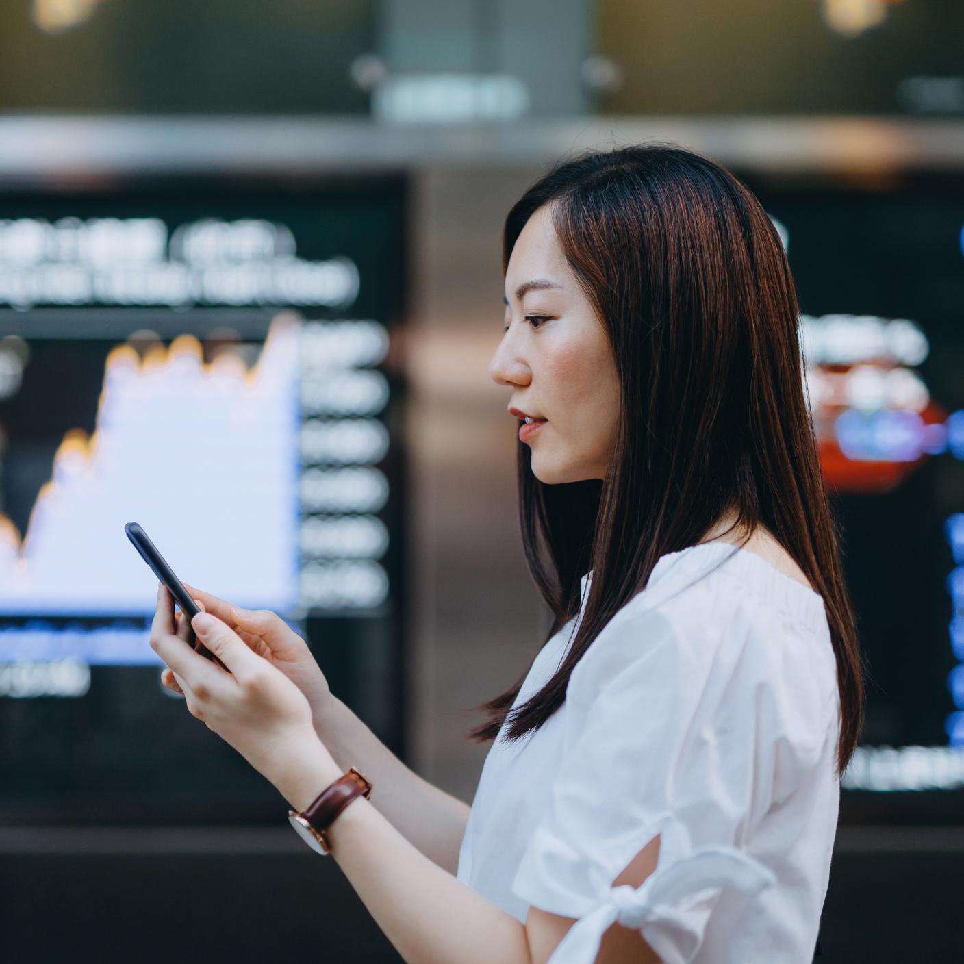 Young Asian businesswoman using smartphone against stock exchange market display screen board