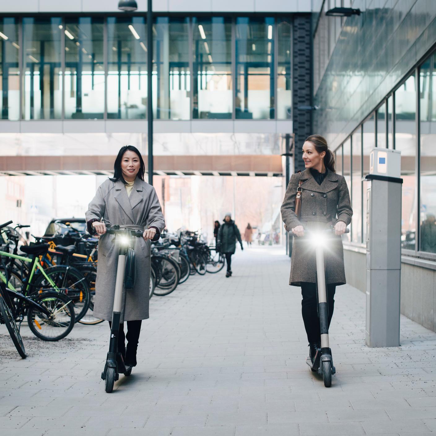 Full length of confident colleagues riding electric push scooter on footpath by building in city