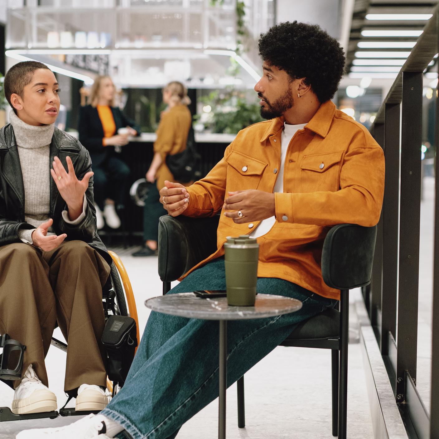 Young person in black leather jacket chats with colleague around coffee table in lobby area