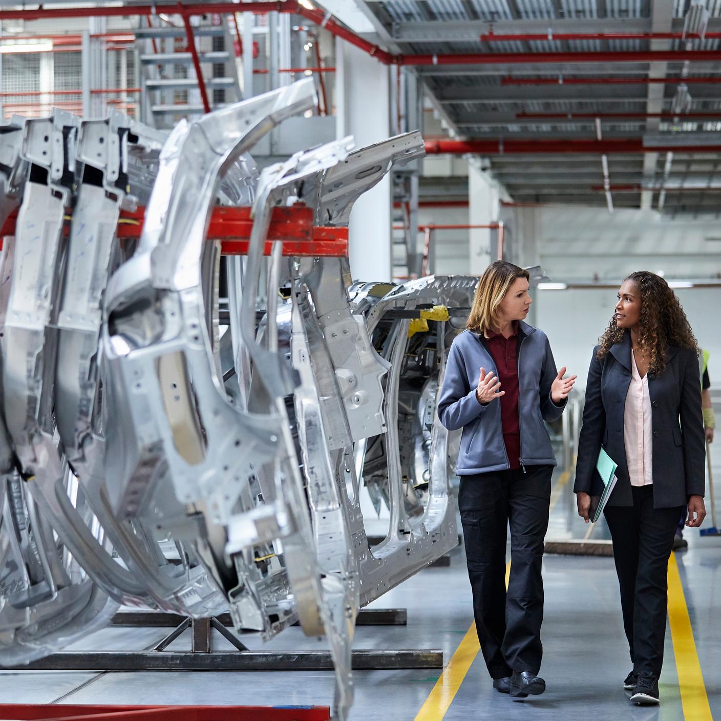 Female engineers discussing by car chassis. Full length of professionals are walking on aisle in factory. Colleagues are in automobile industry.