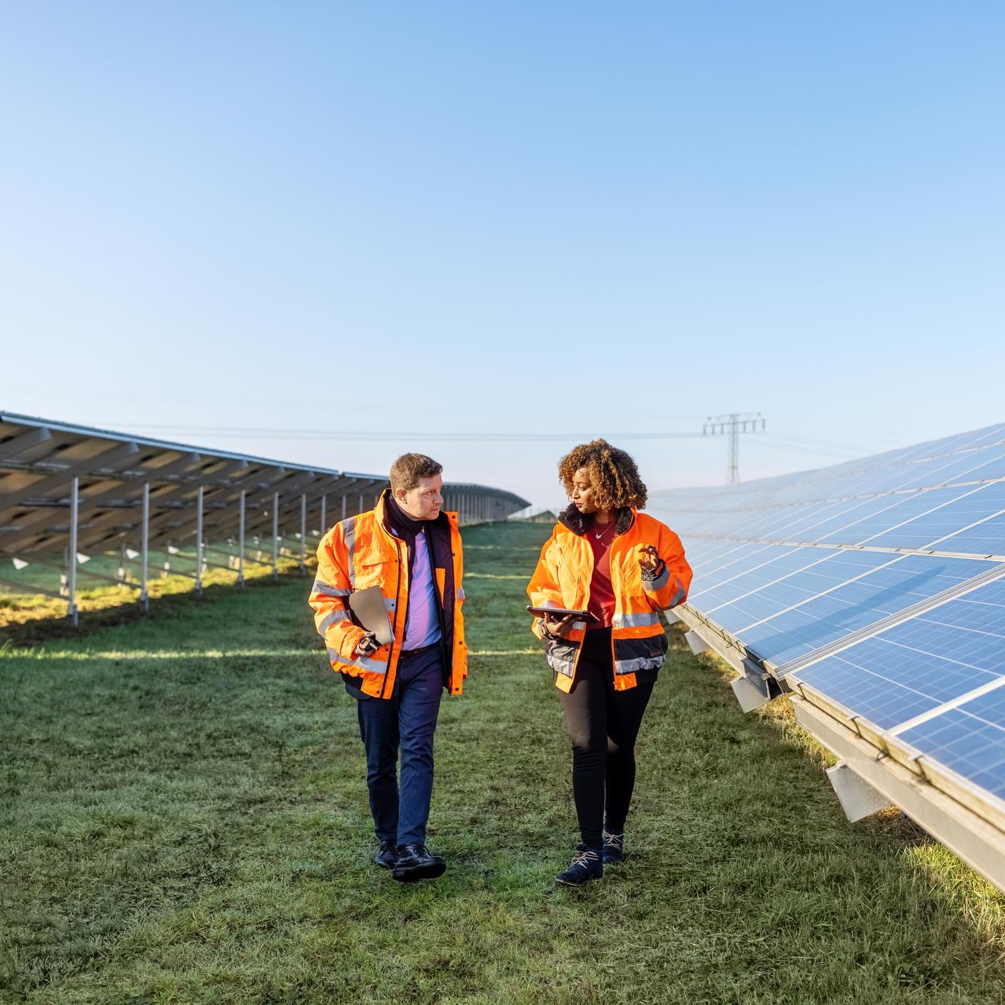 Two technicians in reflective clothing walking between rows of photovoltaic panels at solar farm.