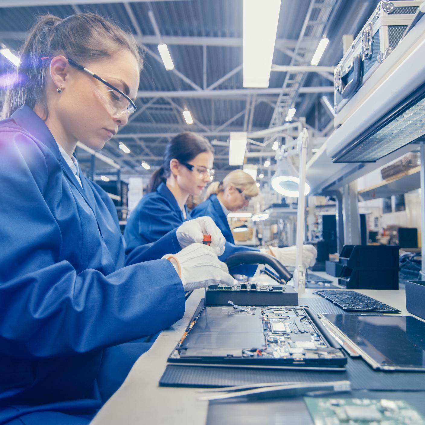 Woman Electronics Factory Worker in Blue Work Coat and Protective Glasses is Assembling Laptop's Motherboard with a Screwdriver.