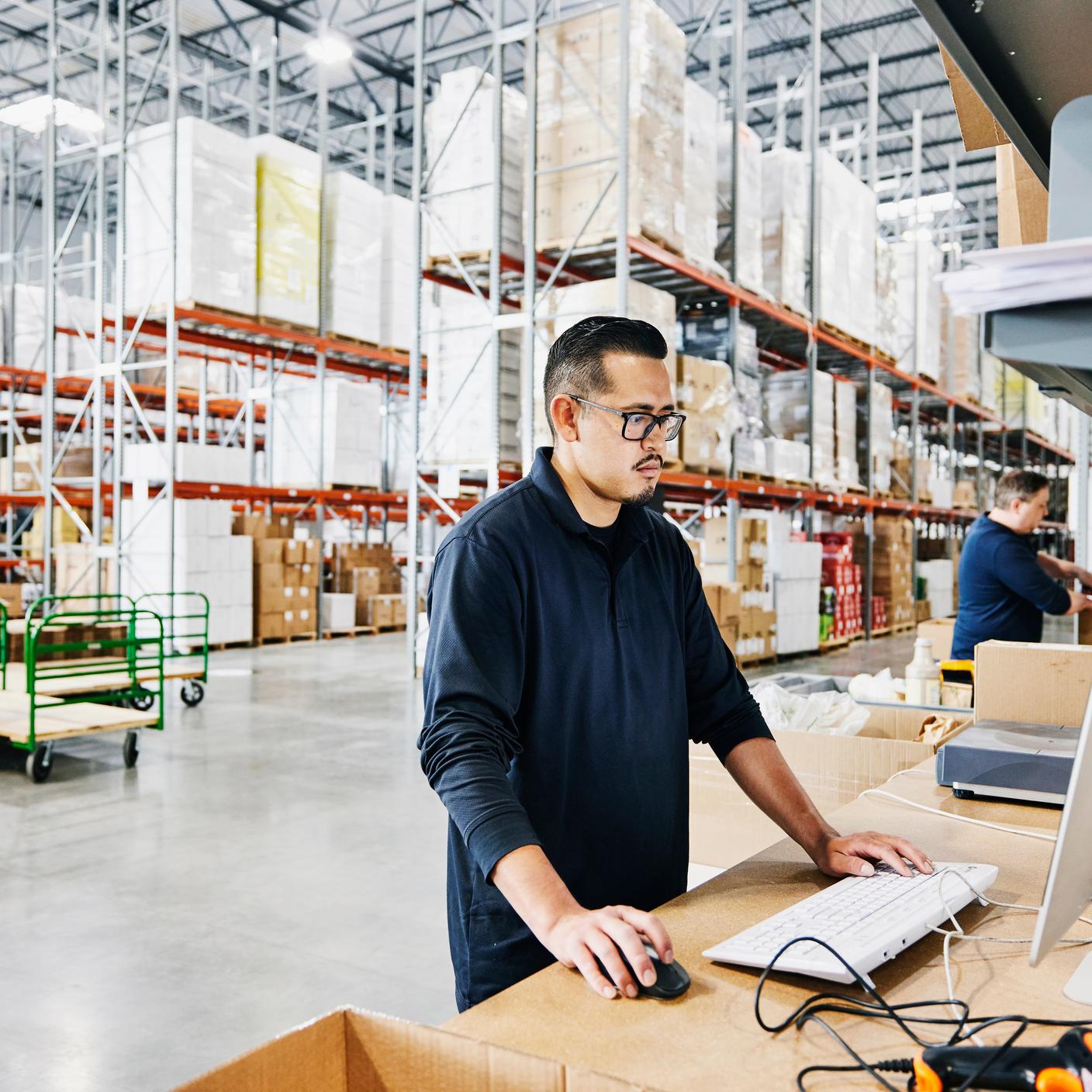 Medium wide shot of male warehouse worker checking orders at computer workstation in warehouse