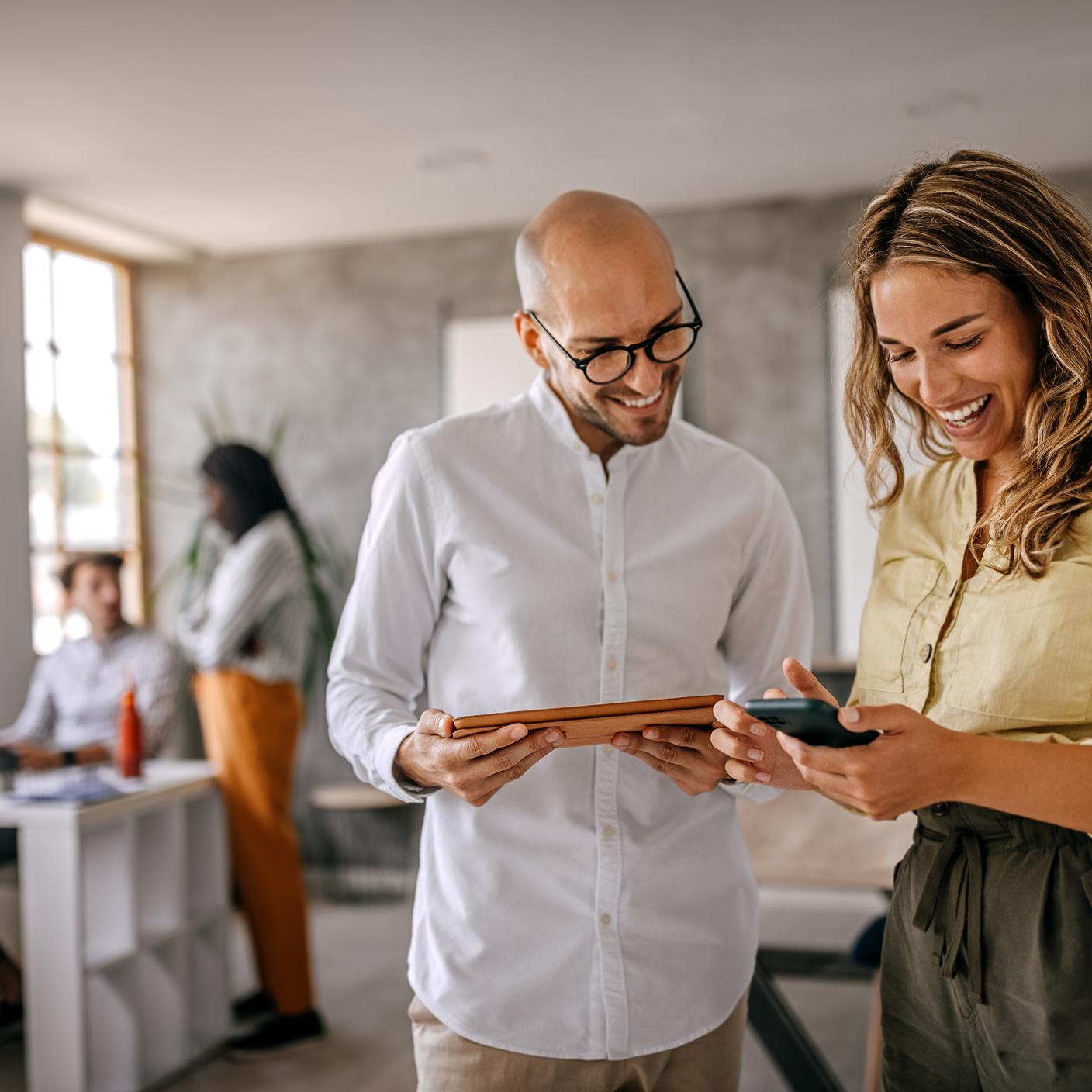 Cheerful and smiling young successful female businesswoman standing with colleague looking at smartphone in modern office and coworking space