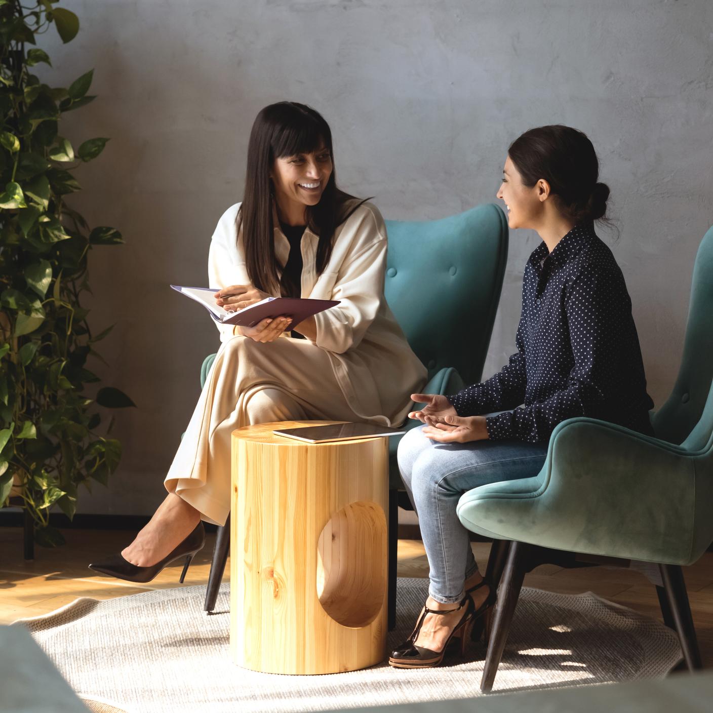 Two friendly women colleagues seated in armchairs enjoy pleasant talk, share information at informal meeting in modern lobby of office. Indian and Caucasian businesswomen conversation during workflow (cropped)