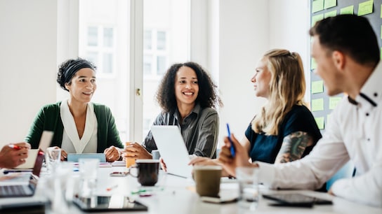 A group of office workers sitting down and talking together at a large desk during a morning meeting.