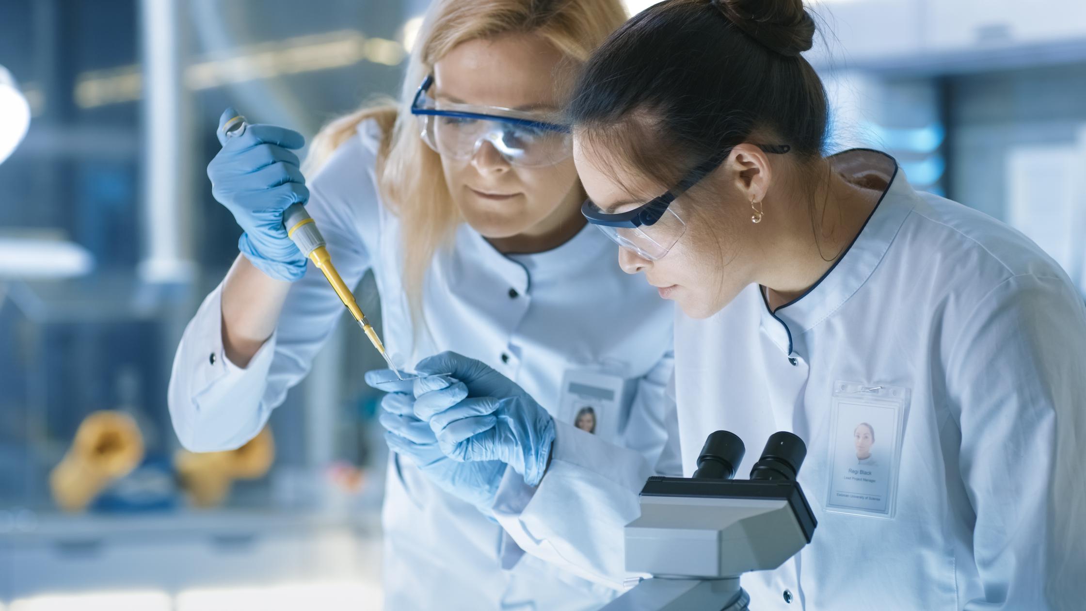Cientista de pesquisa médica deixa cair amostra em lâmina e seu colega a examina sob microscópio. Eles trabalham em um laboratório moderno.