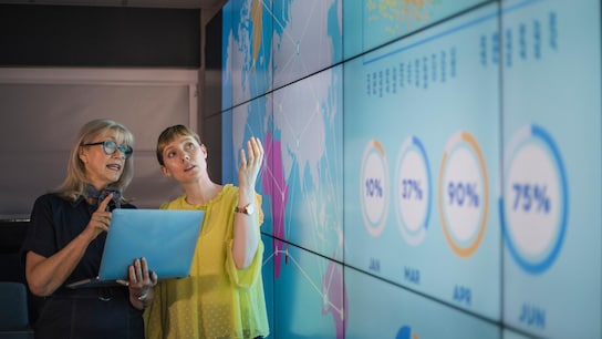 An experienced woman mentors a female colleague, the mature woman is holding a laptop as they debate data from an interactive display; they are both wearing smart casual clothing.